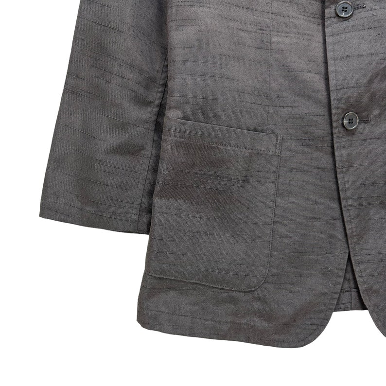 Puede incluir: Una chaqueta gris con un solo bolsillo en el pecho y dos botones. La chaqueta est&aacute; hecha de una tela texturizada.