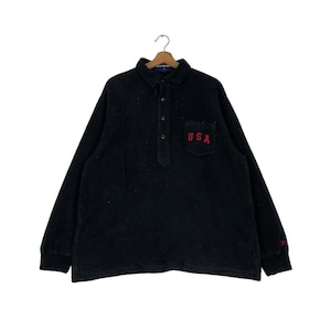 Puede incluir: Camiseta negra de manga larga con cuello abotonado y un pequeño bolsillo en el pecho izquierdo. El bolsillo tiene la palabra "USA" bordada en rojo.