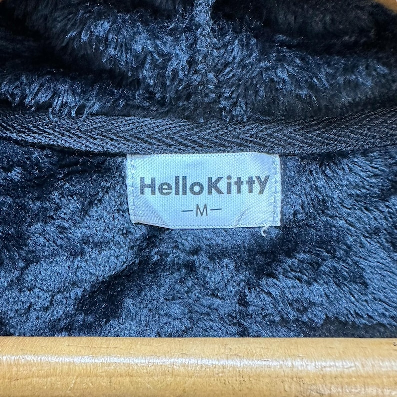Może przedstawiać: Czarna kurtka z kapturem z pluszowym, teksturowanym wnętrzem. Etykieta zawiera napis "Hello Kitty" w białych literach. Kurtka wisi na drewnianym wieszaku.