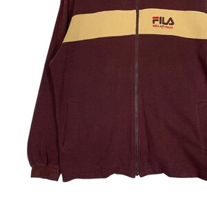 May include: A burgundy zip-up jacket with a tan shoulder panel. The jacket features the FILA logo on the upper right chest. The jacket has two front pockets and a full-length zipper.