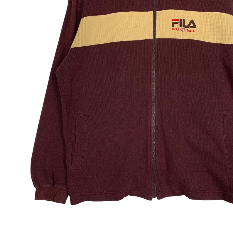 May include: A burgundy zip-up jacket with a tan shoulder panel. The jacket features the FILA logo on the upper right chest. The jacket has two front pockets and a full-length zipper.