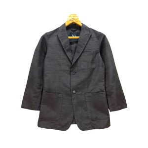 Puede incluir: Una chaqueta de traje gris oscuro con cierre de tres botones y dos bolsillos de parche. La chaqueta tiene una tela texturizada sutil y una etiqueta en forma de V en el lado izquierdo del pecho.