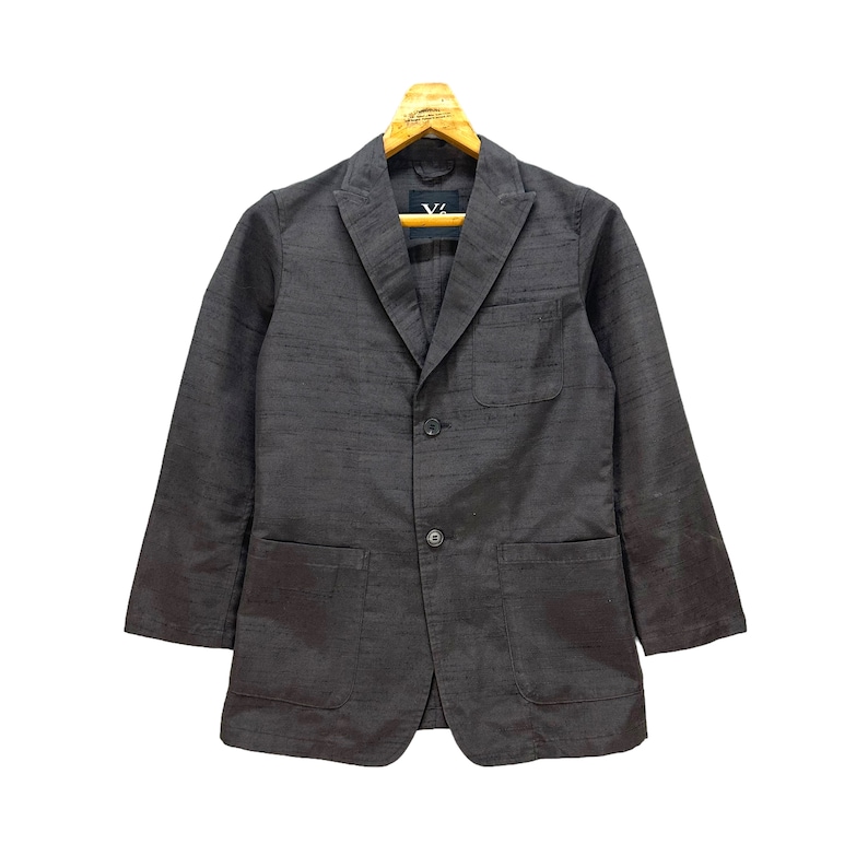 Puede incluir: Una chaqueta de traje gris oscuro con cierre de tres botones y dos bolsillos de parche. La chaqueta tiene una tela texturizada sutil y una etiqueta en forma de V en el lado izquierdo del pecho.