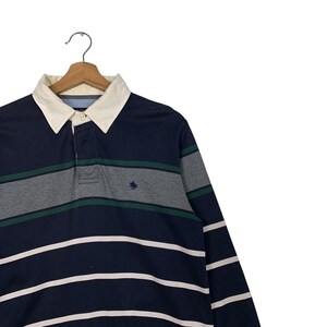 May include: Navy blue long-sleeve polo shirt with white, gray, and green horizontal stripes. The shirt has a cream-colored collar and a small embroidered logo on the chest. The shirt is hanging on a wooden hanger.