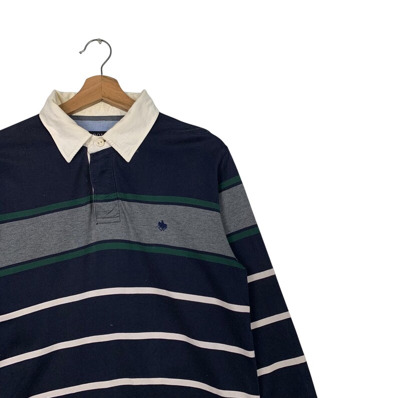 May include: Navy blue long-sleeve polo shirt with white, gray, and green horizontal stripes. The shirt has a cream-colored collar and a small embroidered logo on the chest. The shirt is hanging on a wooden hanger.