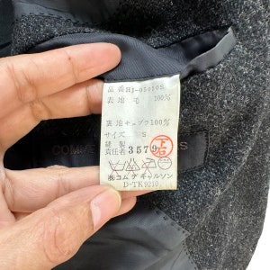 Puede incluir: Una chaqueta de lana negra con una etiqueta blanca que dice "品番 HJ-05050S 表地 毛 100% 裏地 キュプラ 100% サイズ S 縫製 責任著 3579万 (株)コムデギャルソン D-TK 9210".