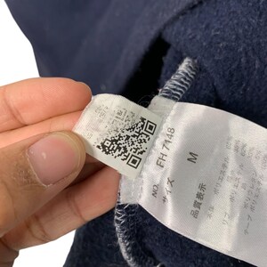 May include: Close-up of a tag attached to a navy blue garment. The tag displays the text "FH 7148" and the size "M". Additional text includes Japanese characters and fabric composition details. A QR code is also visible.