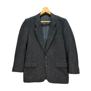 Puede incluir: Una chaqueta de traje gris oscuro con un patr&oacute;n texturizado, colgada de una percha de madera. La chaqueta tiene un cierre de dos botones y dos bolsillos.