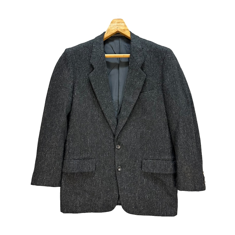 Puede incluir: Una chaqueta de traje gris oscuro con un patr&oacute;n texturizado, colgada de una percha de madera. La chaqueta tiene un cierre de dos botones y dos bolsillos.