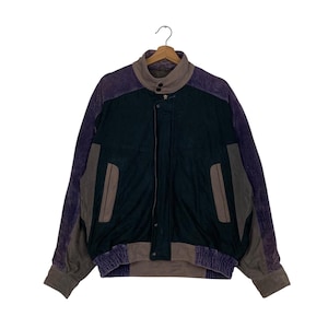 Könnte beinhalten: Eine Vintage-Jacke mit Colorblock-Design. Die Jacke hat eine dunkelgrüne Mittelpartie, ergänzt durch graue und violette Cordärmel und Akzente. Sie hat eine Knopfleiste, einen Kragen und zwei Vordertaschen. Die Jacke hängt an einem Holzkleiderbügel.