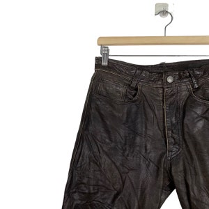 May include: Brown leather pants hanging on a wooden hanger. The pants have a classic five-pocket design and a zipper fly.