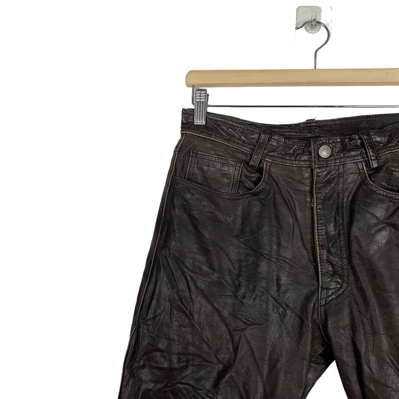 May include: Brown leather pants hanging on a wooden hanger. The pants have a classic five-pocket design and a zipper fly.