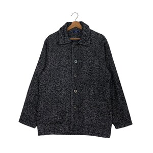 May include: A black and white speckled button-down jacket with a collared neckline and two front pockets. The jacket is suspended on a wooden hanger. The jacket has four buttons down the front and is against a white background.