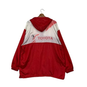 May include: Red and white Toyota windbreaker jacket with a hood. The jacket features the Toyota logo in red and white on the back. The jacket is hanging on a wooden hanger.