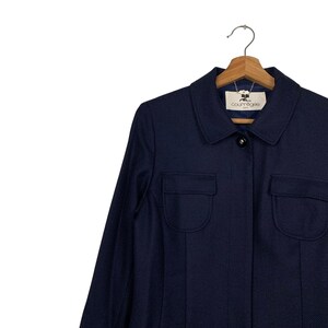 May include: A navy blue jacket with two front pockets and a collared neckline. The jacket has a button closure and the brand name "courr&egrave;ges" is visible on the label. The jacket is hanging on a wooden hanger.