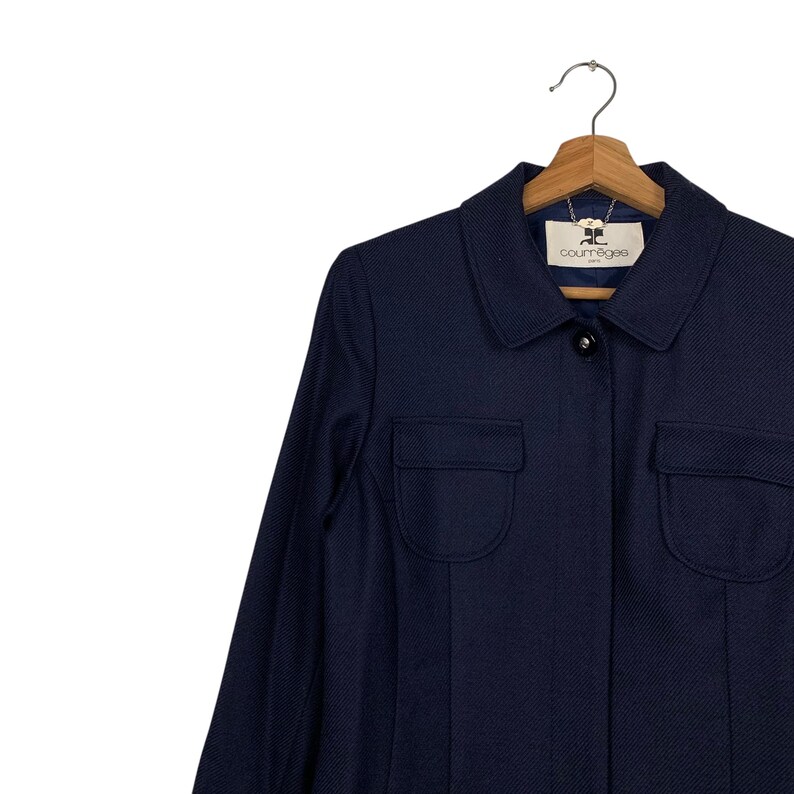 May include: A navy blue jacket with two front pockets and a collared neckline. The jacket has a button closure and the brand name "courr&egrave;ges" is visible on the label. The jacket is hanging on a wooden hanger.