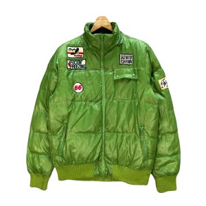 Op de afbeelding: Felgroene pufferjack met meerdere patches en een hoge kraag. Patches met tekst als "Fuckin' Freezing", "Saltwater Fisherman" en het nummer "66". De jas heeft een ritssluiting aan de voorkant en twee zakken.