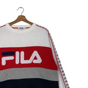 May include: White, red, gray, and navy blue Fila sweatshirt. The sweatshirt features the Fila logo in large letters across the front. The sleeves have a red and white striped design. The sweatshirt is hanging on a wooden hanger.