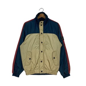 May include: A blue and beige jacket with red trim. The jacket has a zipper closure and a button-up front. It has two pockets on the front and a patch on the left sleeve.