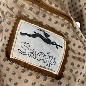 May include: Close-up of a brown suede jacket interior. The label reads "Sacip MADE IN ITALY" with a black leaping animal logo. The lining is a tan fabric with a repeating pattern of small gray squares.