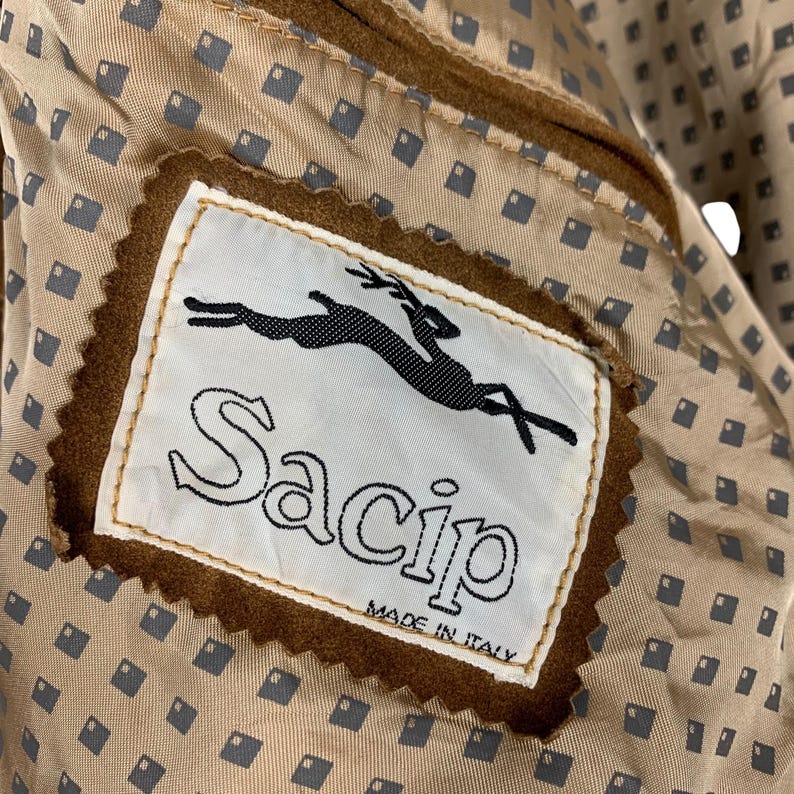 May include: Close-up of a brown suede jacket interior. The label reads "Sacip MADE IN ITALY" with a black leaping animal logo. The lining is a tan fabric with a repeating pattern of small gray squares.