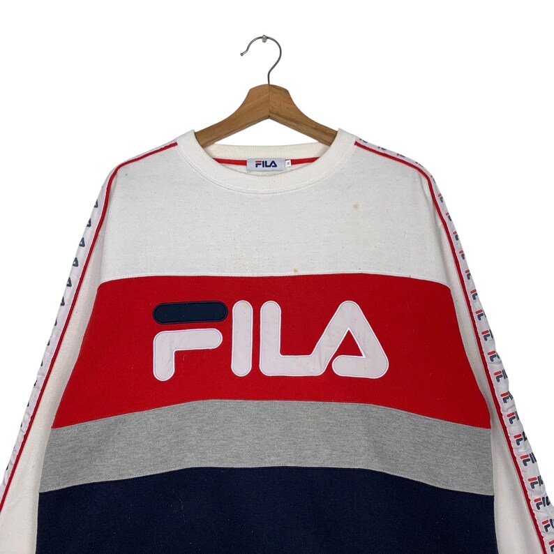 May include: A color-blocked Fila sweatshirt in white, red, gray, and navy blue. The Fila logo is prominently displayed in white across the chest. The sleeves have a red and white stripe design with the Fila logo. The sweatshirt is hanging on a wooden hanger.
