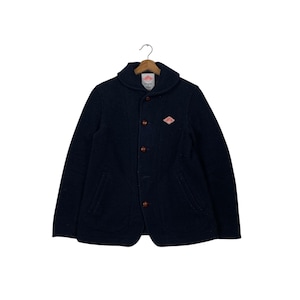 Puede incluir: Chaqueta azul marino con cuello chal y cuatro botones marrones. La chaqueta tiene dos bolsillos delanteros y un pequeño parche con el logotipo rojo y naranja en el pecho. La chaqueta está colgada de una percha de madera.