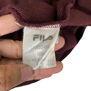 May include: Close-up of a maroon garment tag with the FILA logo and text. The tag includes the text "15JDU3456" and other Japanese text. The garment is a deep maroon color.
