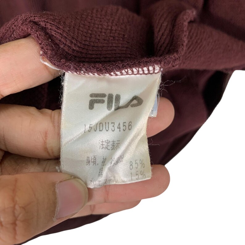 May include: Close-up of a maroon garment tag with the FILA logo and text. The tag includes the text "15JDU3456" and other Japanese text. The garment is a deep maroon color.