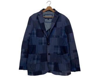 Op de afbeelding: Een blauwe patchwork blazer met een sluiting met twee knopen, twee voorzakken en een revers met inkeping. De jas heeft een verscheidenheid aan blauwe stofpatronen, waaronder ruit- en streepontwerpen. De blazer hangt aan een houten hanger.