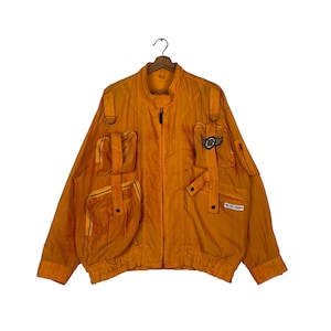 Puede incluir: Una chaqueta naranja brillante con múltiples bolsillos y cierre de cremallera. La chaqueta tiene un tirador de cremallera negro y un parche negro con el texto "KC THE MONKEY" en el lado izquierdo.
