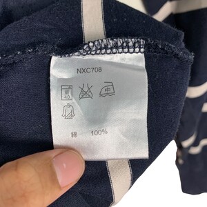 May include: Close-up of a navy blue garment tag with the text "NXC708". The tag displays laundry symbols and the text "綿 100%". The garment has white stripes.