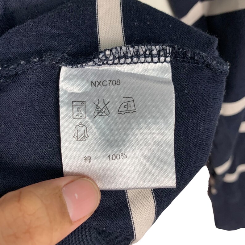 May include: Close-up of a navy blue garment tag with the text "NXC708". The tag displays laundry symbols and the text "綿 100%". The garment has white stripes.