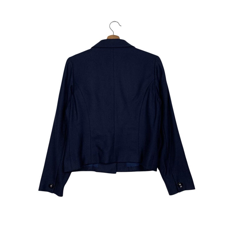 May include: A navy blue blazer, viewed from the back, hanging on a wooden hanger. The blazer has a classic design with a notched lapel, two buttons on each cuff, and a single-button closure. The fabric appears to be a smooth, woven material.