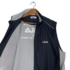 Puede incluir: Una chaqueta negra y gris sin mangas con cierre de cremallera. La chaqueta tiene un forro de malla y el logotipo de Fila est&aacute; bordado en la parte delantera.