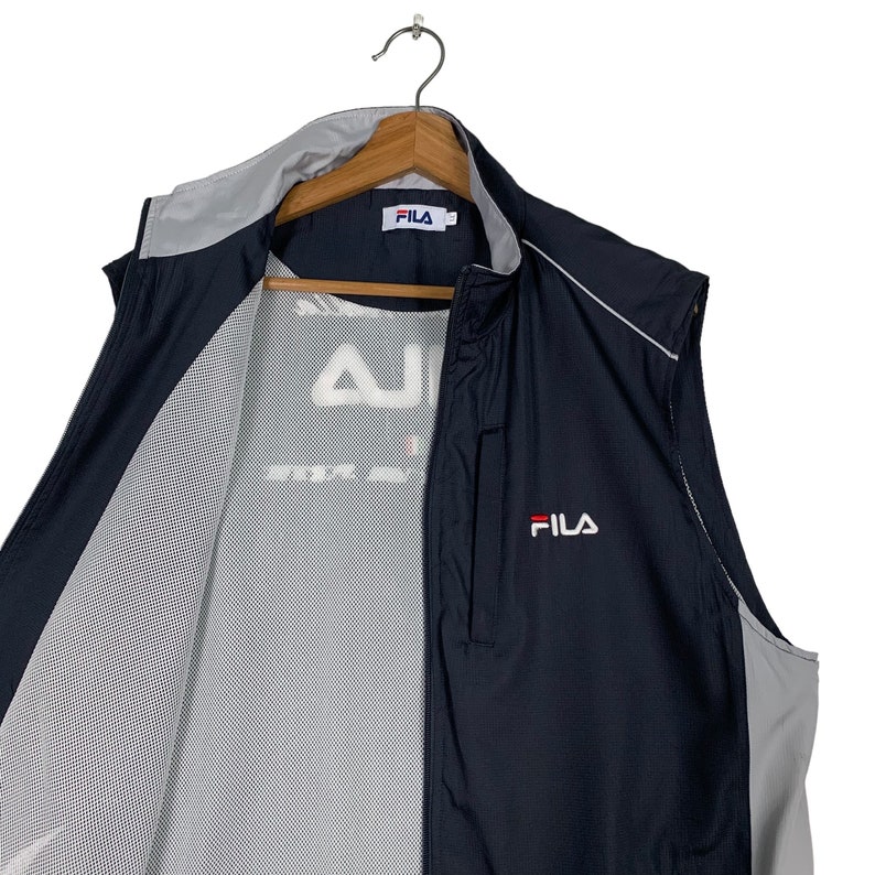 Puede incluir: Una chaqueta negra y gris sin mangas con cierre de cremallera. La chaqueta tiene un forro de malla y el logotipo de Fila est&aacute; bordado en la parte delantera.