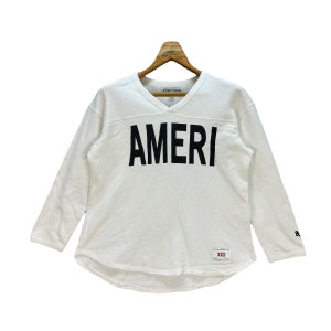 May include: White long-sleeved shirt with a v-neck and the word "AMERI" printed in black on the front. The shirt has a tag that says "Americana" and "XXS".