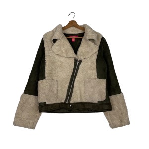 Puede incluir: Una chaqueta elegante con una mezcla de texturas y colores. La chaqueta presenta un material crema, esponjoso, similar a la piel de oveja en el cuello, los paneles delanteros y los puños, contrastado con mangas y parte inferior del cuerpo de ante verde oscuro. Tiene cierre de cremallera diagonal y dos bolsillos.