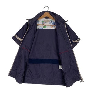 May include: A navy blue short-sleeved jacket with a zip closure and a white lining featuring a palm tree and boat print. The jacket has a red and white trim and a zipped pocket on the left side.