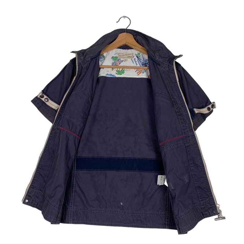 May include: A navy blue short-sleeved jacket with a zip closure and a white lining featuring a palm tree and boat print. The jacket has a red and white trim and a zipped pocket on the left side.