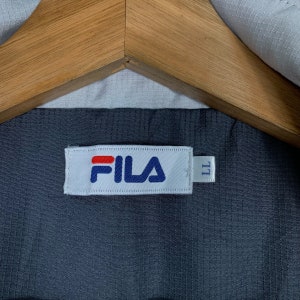 Puede incluir: Una etiqueta blanca con la palabra "FILA" en letras azules y rojas. La etiqueta est&aacute; unida a una tela gris oscura.