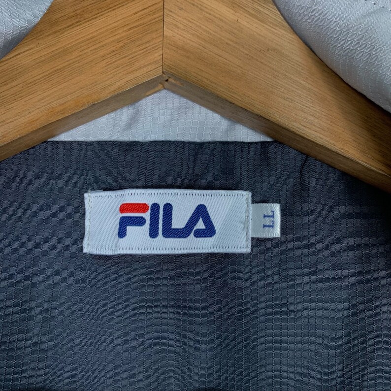 Puede incluir: Una etiqueta blanca con la palabra "FILA" en letras azules y rojas. La etiqueta est&aacute; unida a una tela gris oscura.