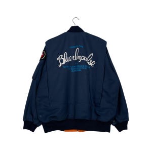 Peut inclure: Une veste bomber bleu foncé avec un patch sur la manche gauche et le texte "Blue Impulse" au dos. Le texte est en blanc, bleu et bleu clair. La veste a une fermeture éclair et deux poches sur le devant.