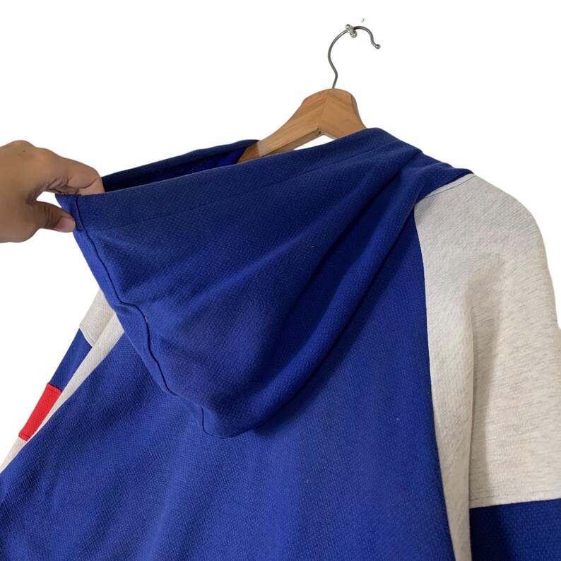 Puede incluir: Una sudadera con capucha azul, blanca y gris con cierre de cremallera. La capucha est&aacute; doblada hacia abajo y la sudadera est&aacute; colgada de una percha de madera.
