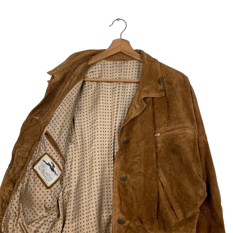 May include: A brown suede jacket with a button-up front, a collared neckline, and a pocket on the front. The jacket is hanging on a wooden hanger. The interior lining is a light tan color with a small square pattern. The brand label "Sacip" is visible.