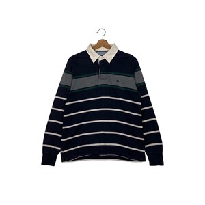 May include: A long-sleeved rugby shirt with navy blue and white horizontal stripes. The shirt features a white collar, a three-button placket, and gray and green horizontal stripes across the chest. A small emblem is visible on the chest.