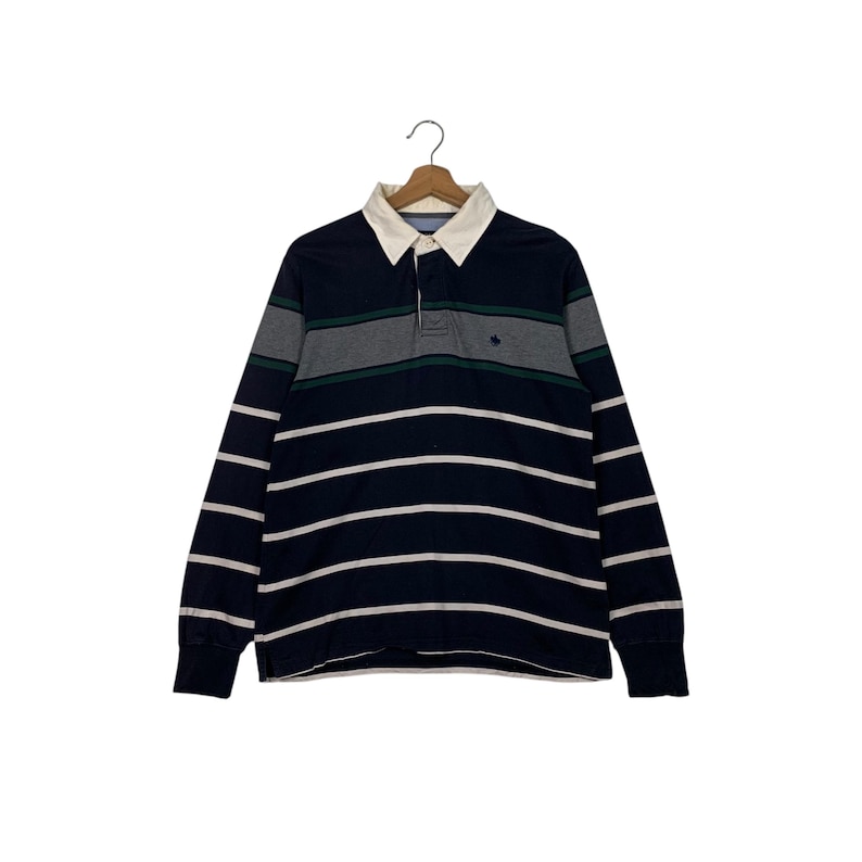 May include: A long-sleeved rugby shirt with navy blue and white horizontal stripes. The shirt features a white collar, a three-button placket, and gray and green horizontal stripes across the chest. A small emblem is visible on the chest.