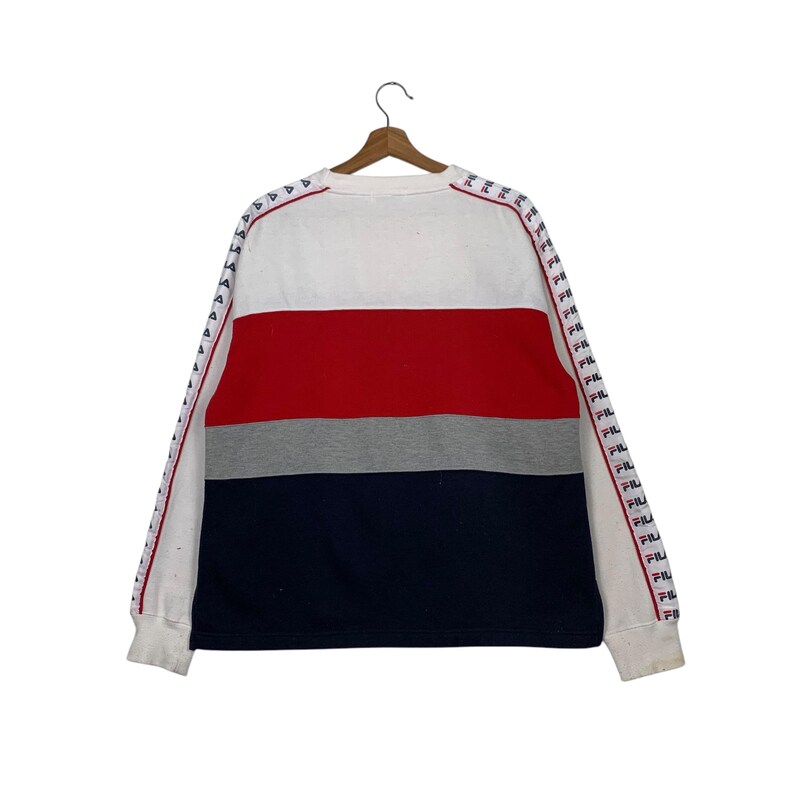 May include: A white, red, gray, and navy blue color-blocked sweatshirt. The long sleeves have a white stripe with red triangles and the word "FILA" printed repeatedly. The sweatshirt is hanging on a wooden hanger.