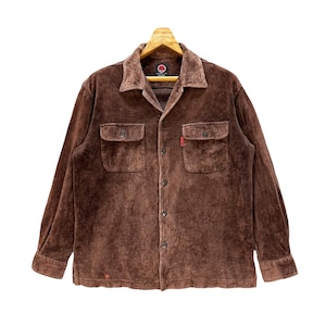 May include: A brown corduroy long-sleeved shirt with a button-up front and two flap pockets. The shirt has a red circle logo on the left side of the collar.