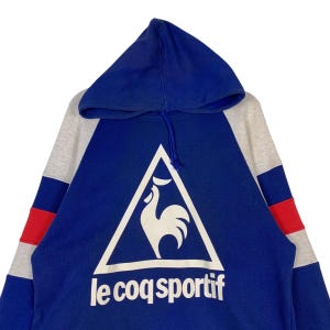 Puede incluir: Una sudadera con capucha azul, gris y roja con un logotipo de gallo blanco y el texto "le coq sportif" en la parte delantera.